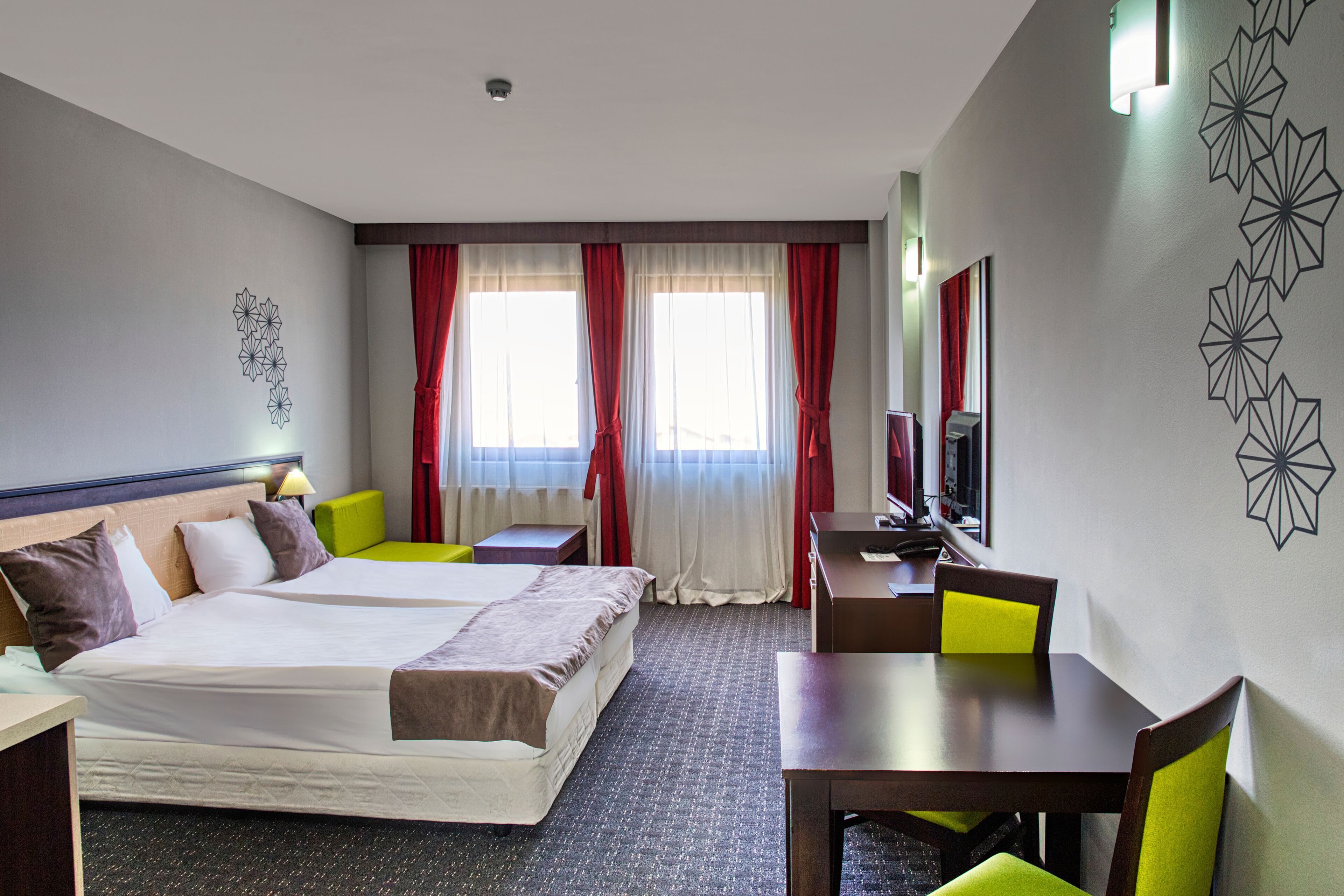 Une chambre d’hôtel comprenant un lit, deux tables de chevet, un bureau avec une chaise, une télévision et une fenêtre avec des rideaux.