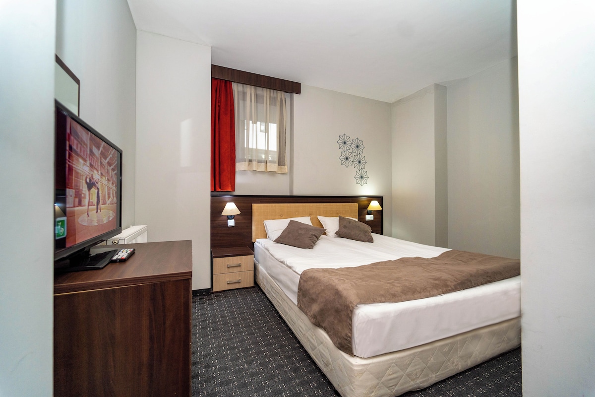 A hotel room with a bed, bedside tables, a TV, and a window with curtains.