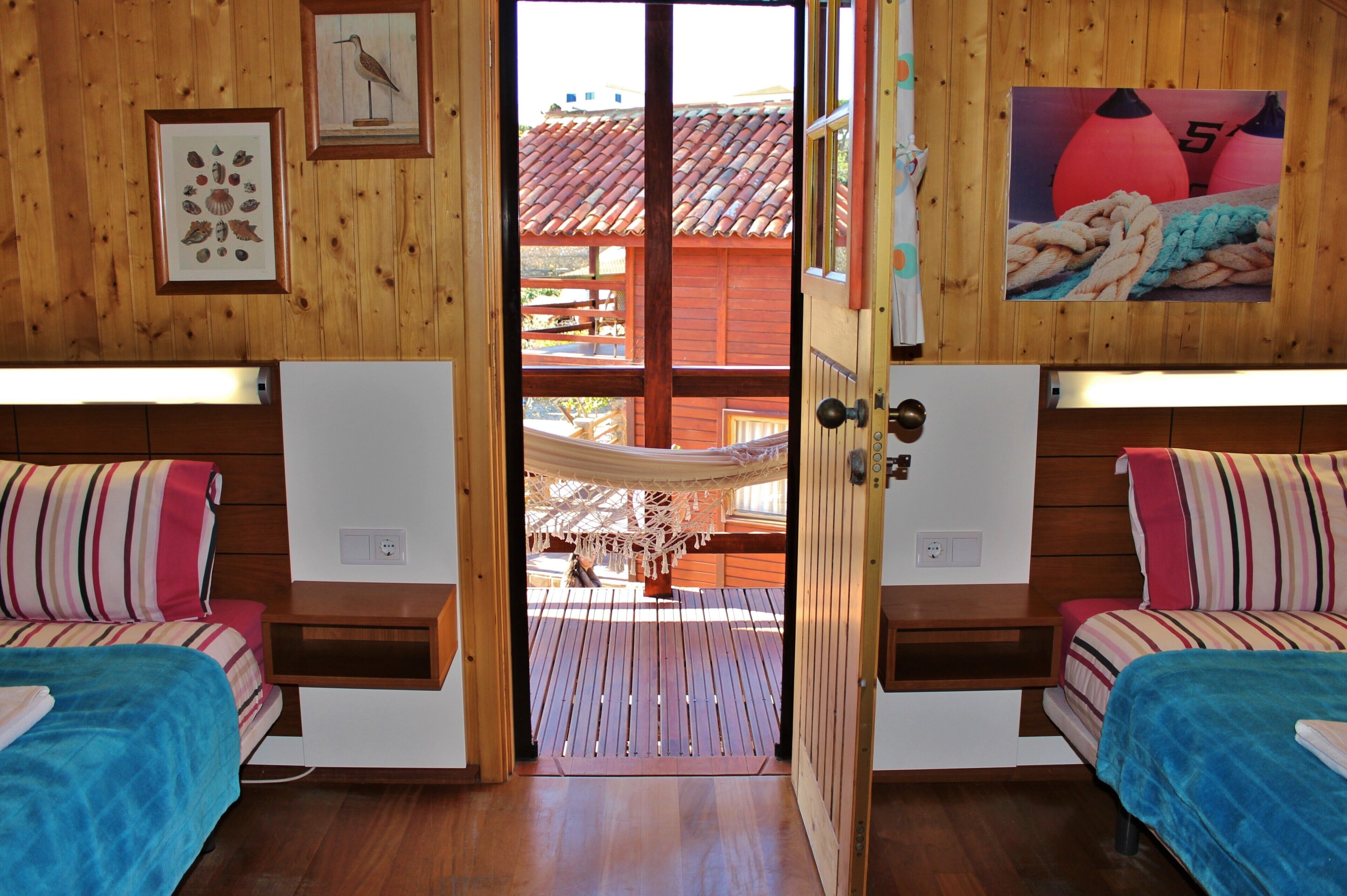 Une chambre avec deux lits, des murs en bois, un hamac et une vue sur un bâtiment aux toits rouges situé à l’extérieur.