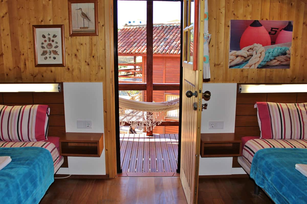 Une chambre avec deux lits, des murs en bois, un hamac et une vue sur un bâtiment aux toits rouges situé à l’extérieur.
