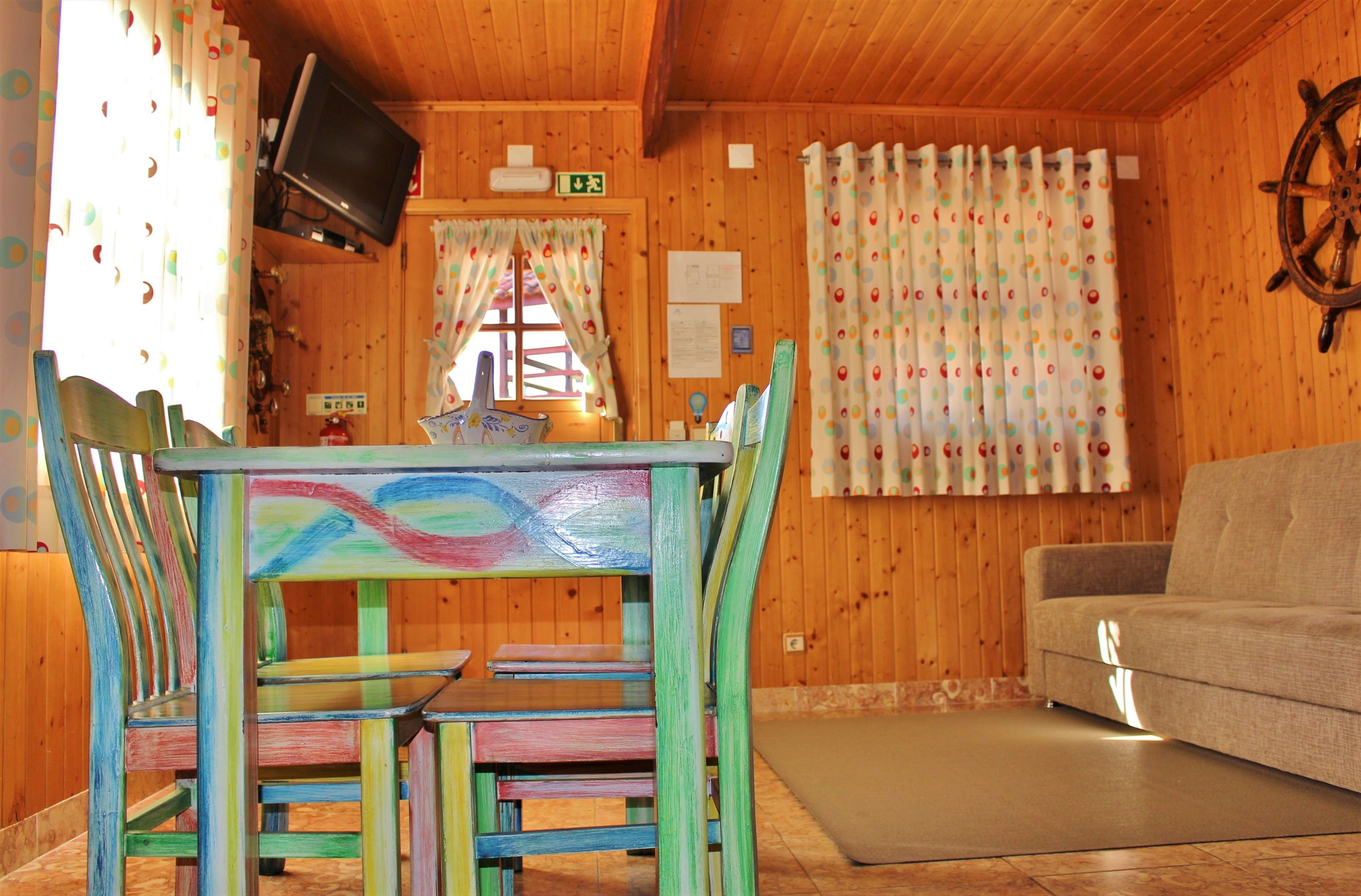 Een houten blokhut interieur met een eettafel, kleurrijke stoelen, een bank en een aan de muur bevestigde televisie.
