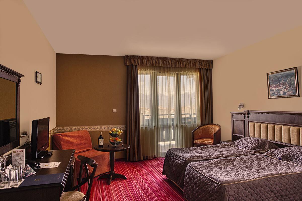 A hotel room with two beds, a desk with a chair, a small table with a fruit bowl, a television, and a window with curtains.