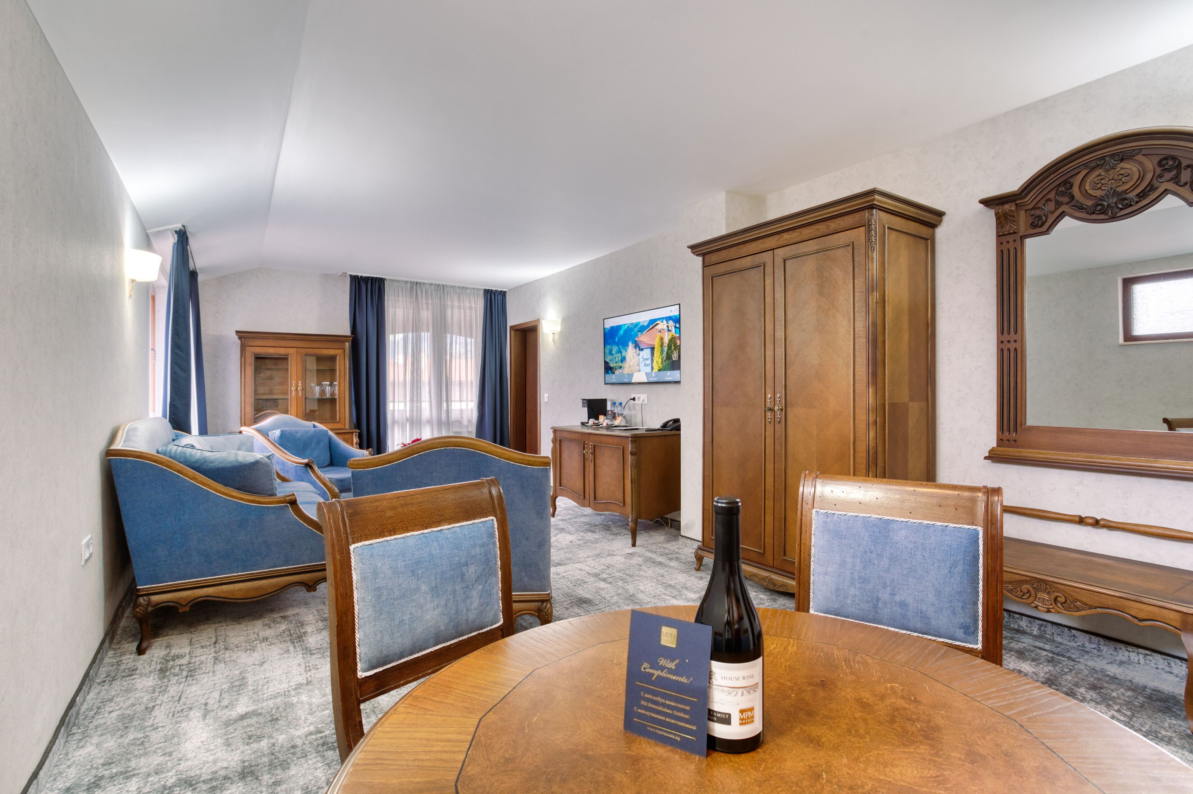 A hotel room with a dining table, two blue upholstered chairs, a wooden cabinet, a television, and a large mirror.