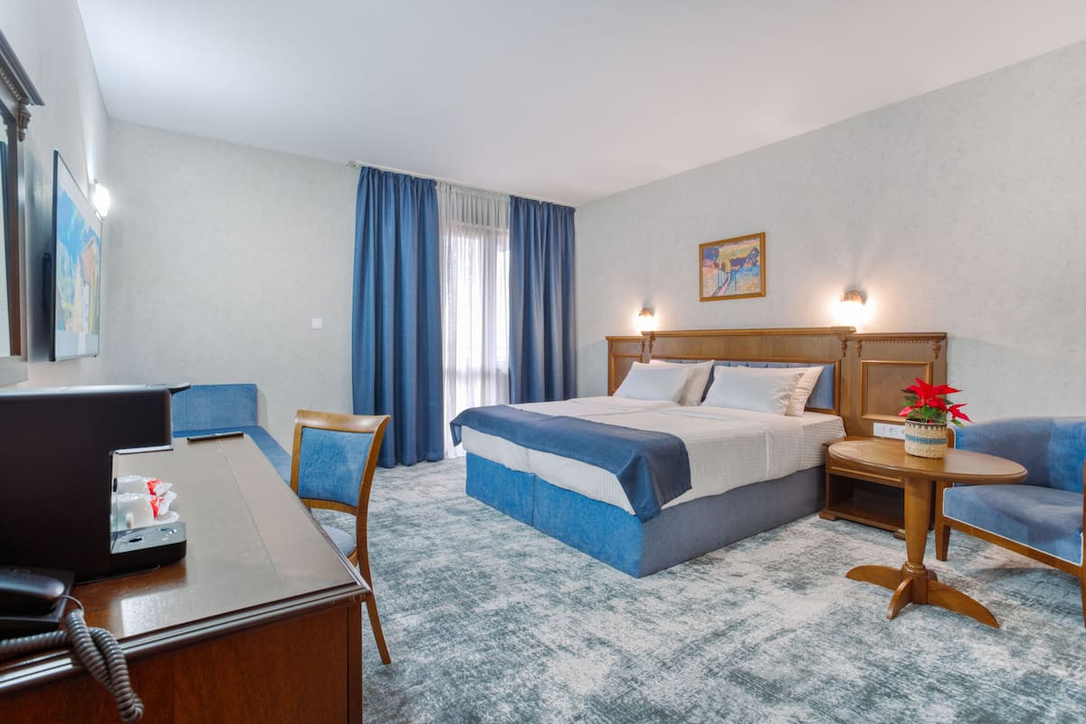 A hotel room with a bed, a desk, a chair, a small table, and a potted plant.