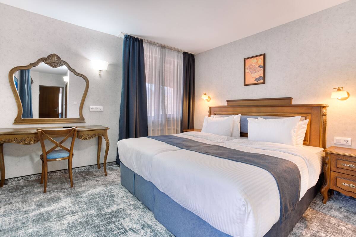 A hotel room with a large bed, a desk with a chair, a mirror, and a nightstand with a lamp.
