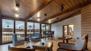 A spacious indoor area with wooden ceiling beams, large windows, and a view of a ski lift. There are several seating areas with tables and chairs. - MPM Sport Hotel (Bansko)