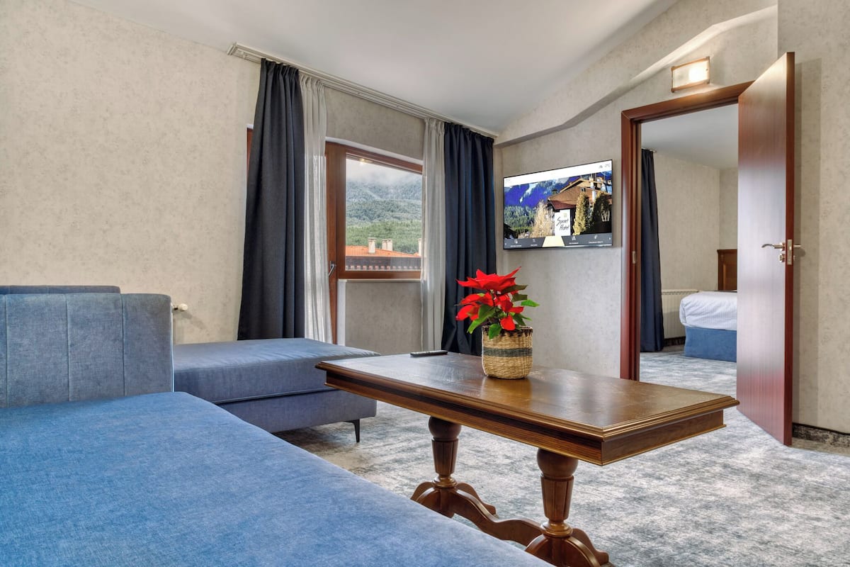 A hotel room with a sofa, a small table with a potted plant, a bed, and a view of mountains through the window.