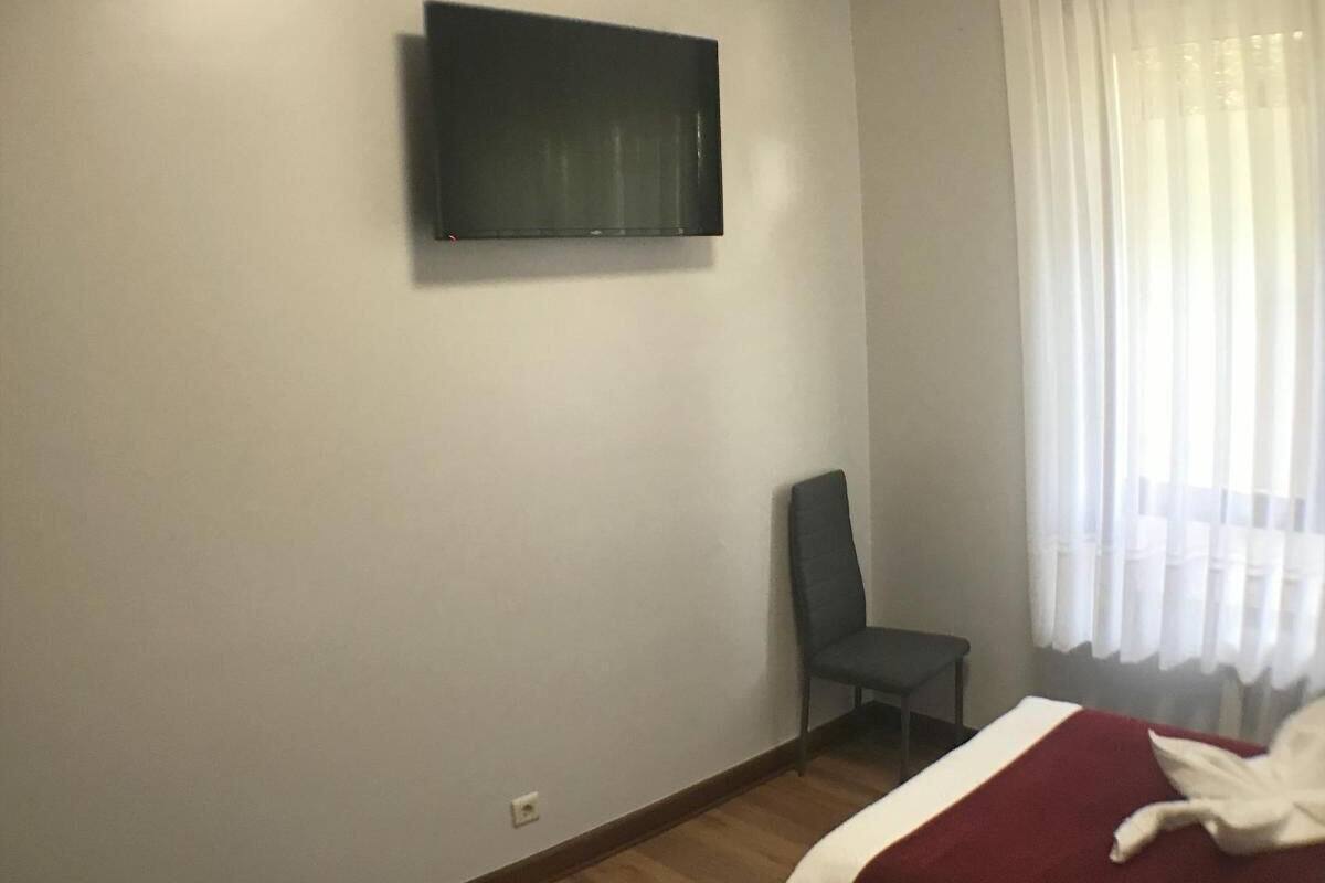 Une pièce comprenant un lit, une chaise, un téléviseur fixé au mur et une fenêtre avec des rideaux.