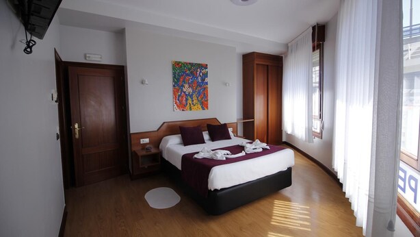 A hotel room with a wooden floor, a bed with a white and maroon bedspread, a colorful abstract painting on the wall, and a window with white curtains. - Pensión La Salve (Bilbao)