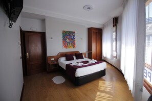 A hotel room with a wooden floor, a bed with a white and maroon bedspread, a colorful abstract painting on the wall, and a window with white curtains. - Pensión La Salve (Bilbao)