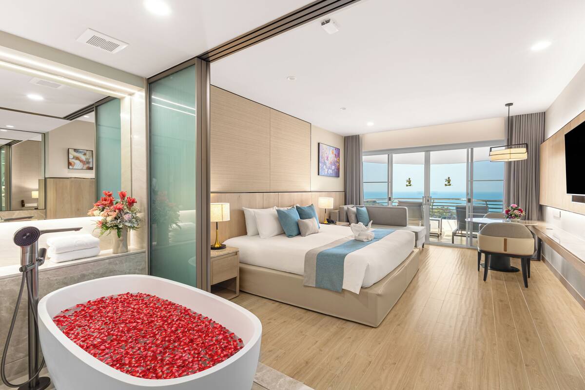 A modern hotel room with a large bed, a bathtub filled with red flowers, a balcony with a view, and a dining area.