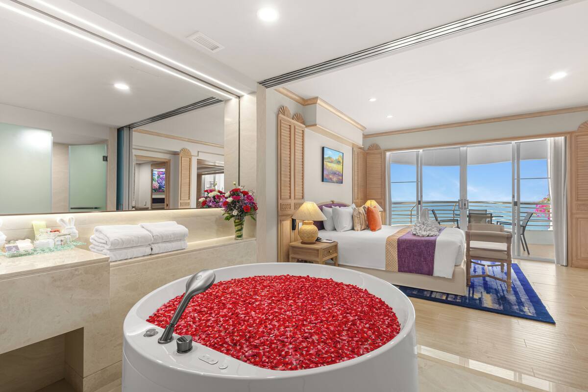 A hotel room with a large bathtub filled with rose petals, a bed with a view of the sea, and a balcony with a view of the ocean.