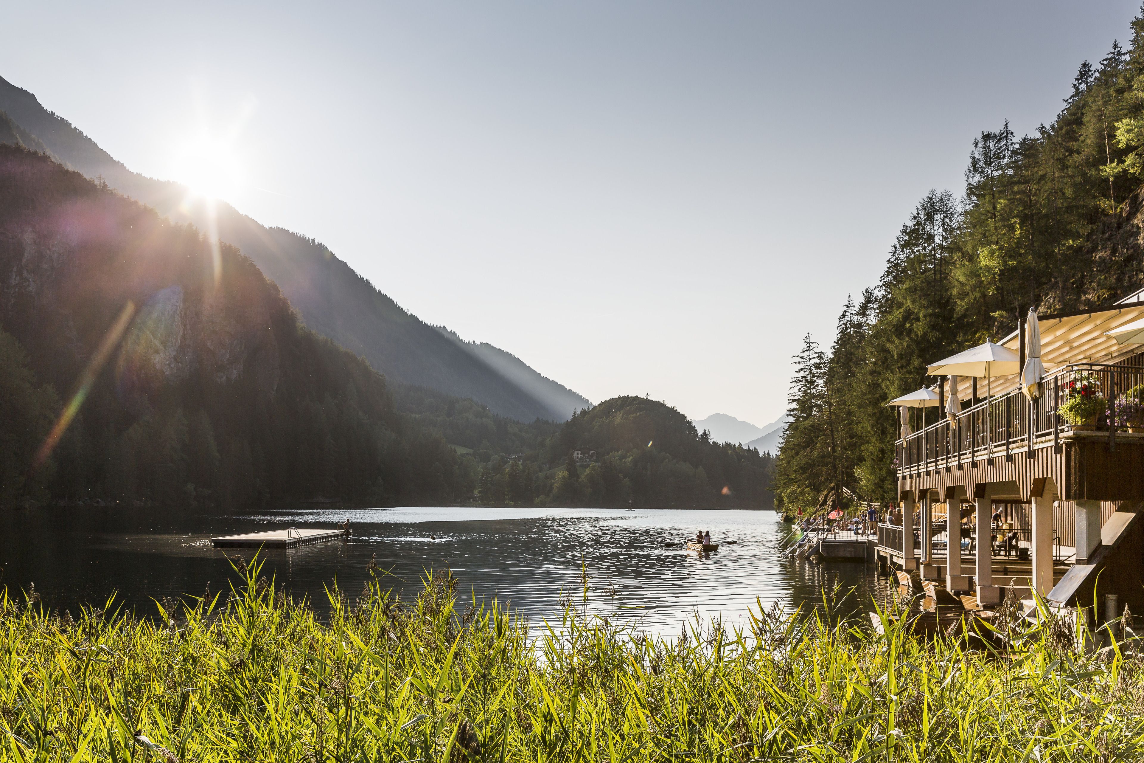 Une vue sur le lac avec un quai en bois, un bâtiment doté d’un balcon surplombant l’eau et un paysage montagneux en arrière-plan.