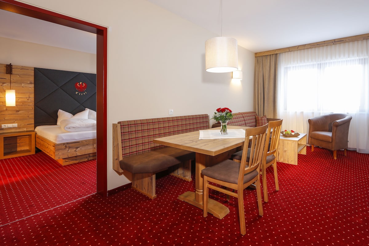 Ein Hotelzimmer mit Bett, Holz-Kopfstück, einer Bank, einem Tisch mit Stühlen, einer Vase mit Blumen und einem Fenster mit Vorhängen.