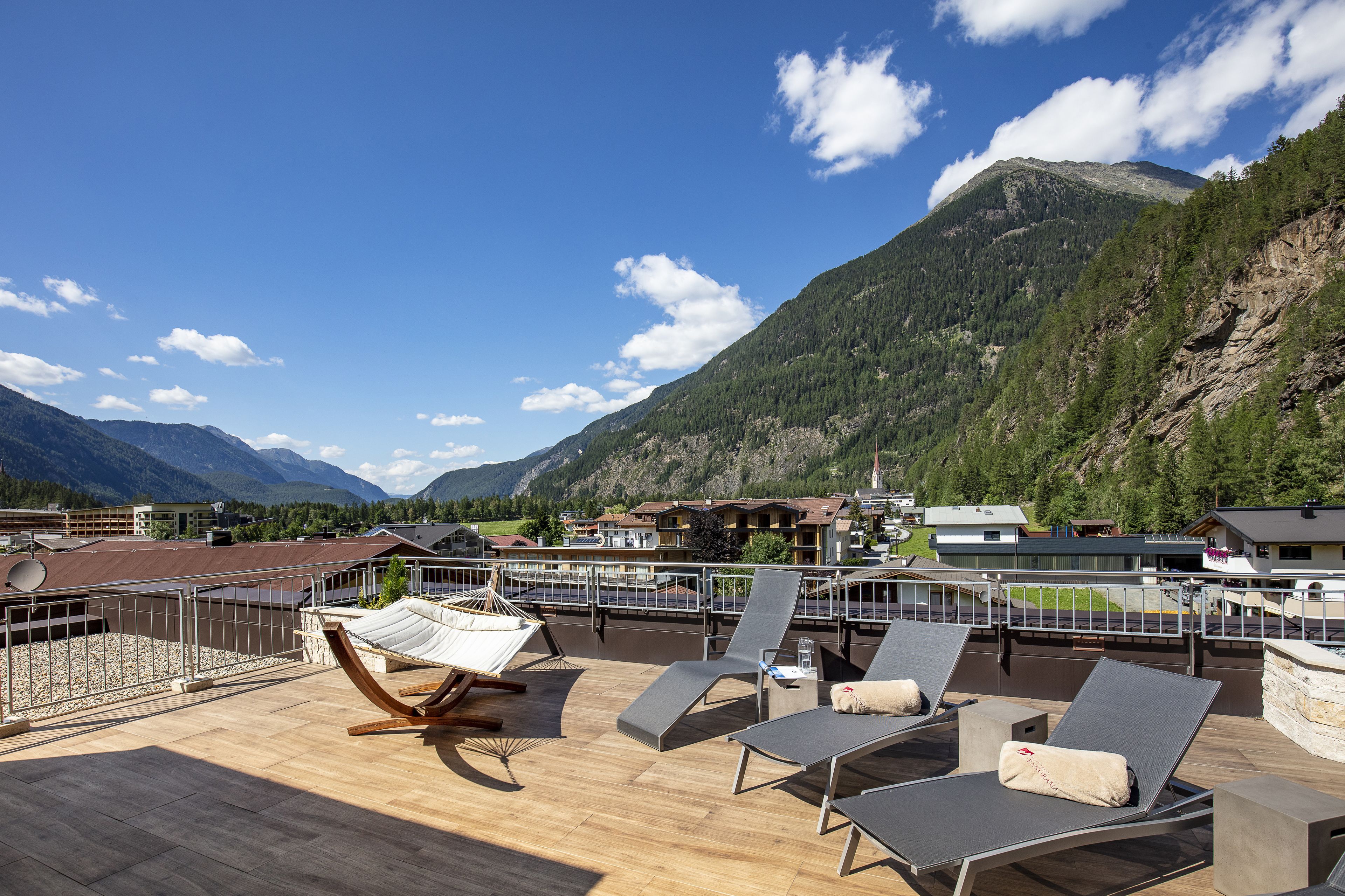 Une terrasse dotée de chaises longues et d’un hamac, offrant une vue sur un paysage montagneux avec des bâtiments en arrière-plan.