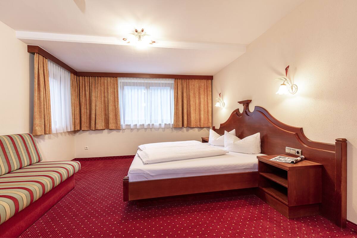 Ein Hotelzimmer mit Bett, Sofa, Nachttisch, Lampe und einem Fenster mit Vorhängen.
