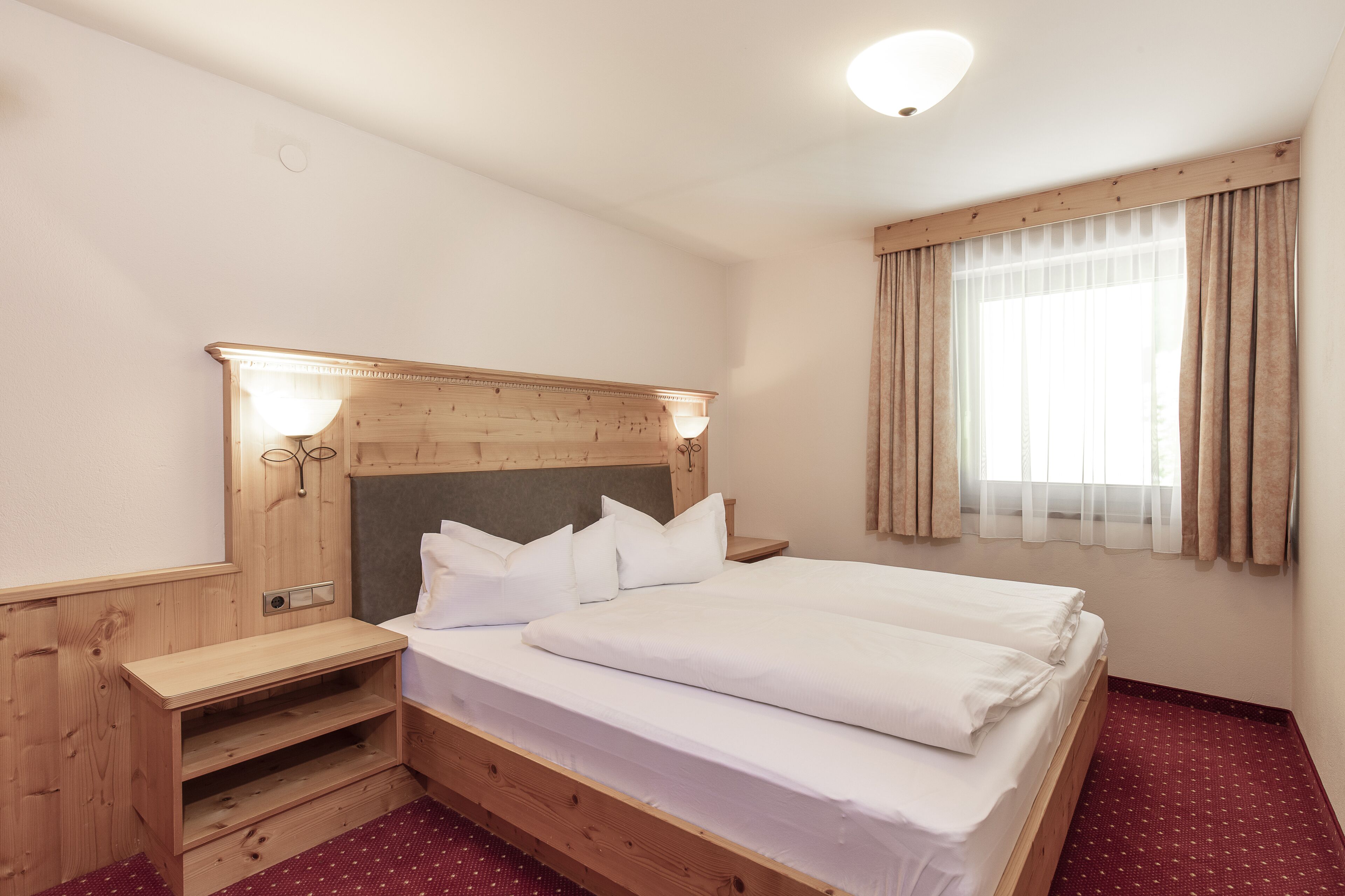 A neatly made bed with a wooden headboard and bedside tables, a window with curtains, and a red carpeted floor.