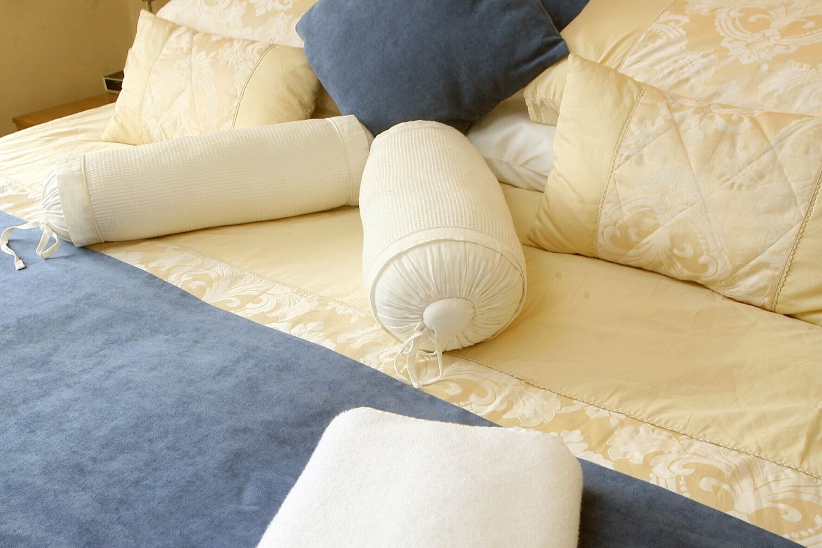 Una cama bien tendida con un cabecero azul, almohadas blancas y estampadas, y una toalla blanca doblada.