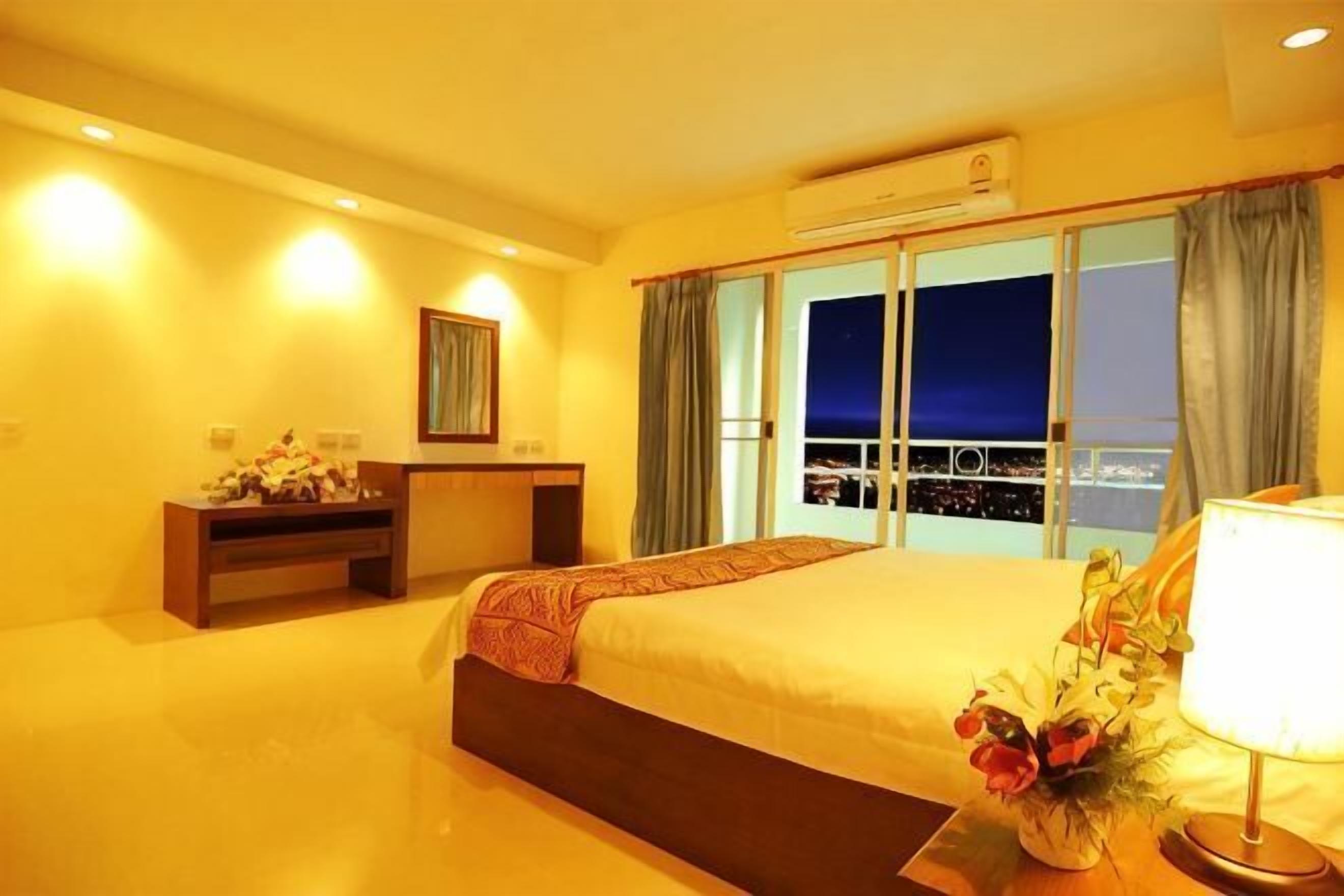 A hotel room with a large bed, a desk, a chair, a nightstand, a lamp, and a view of the city at night.