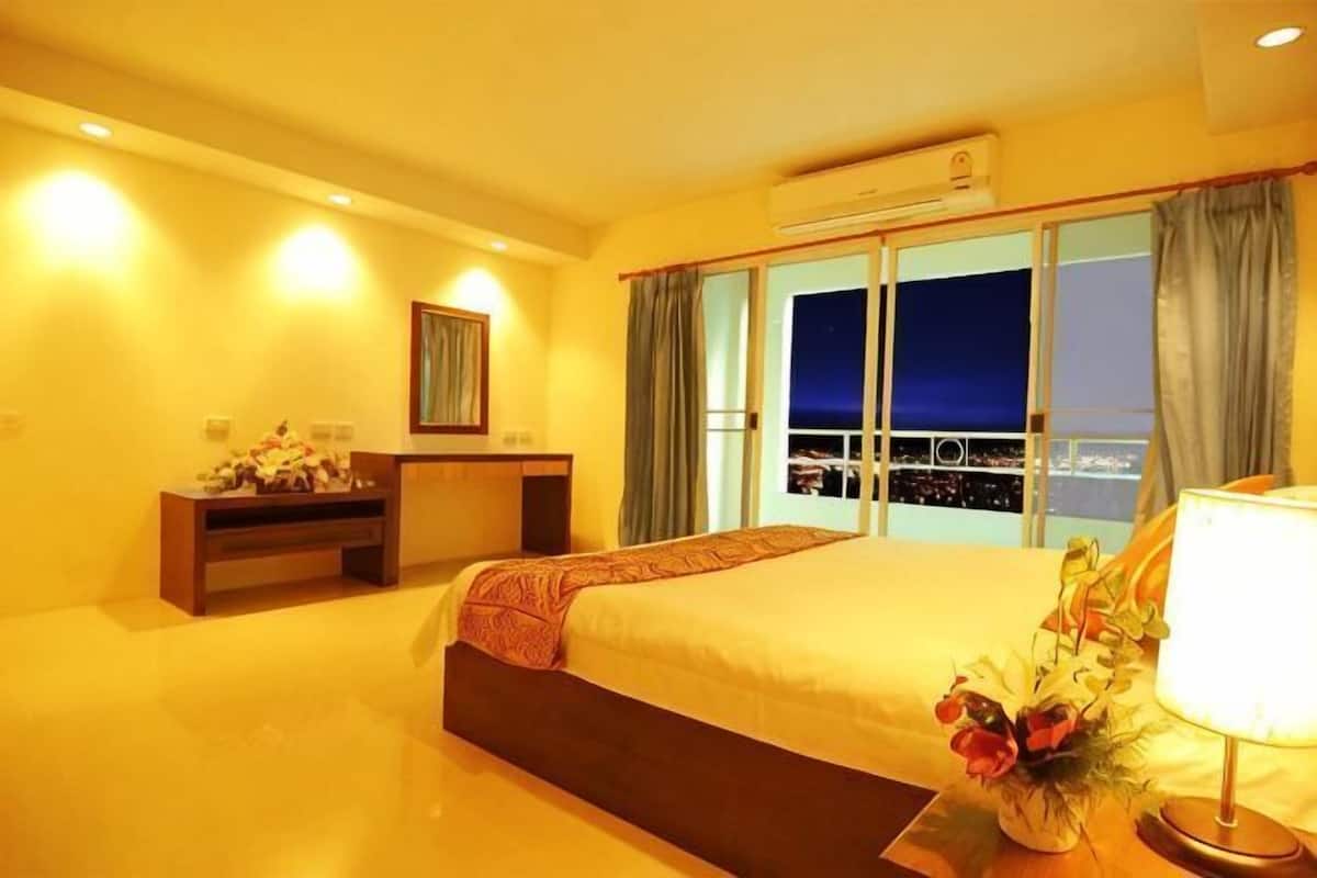 Quarto de hotel com uma cama grande, uma escrivaninha, uma cadeira, uma mesa de cabeceira, um abajur e vista para a cidade à noite.