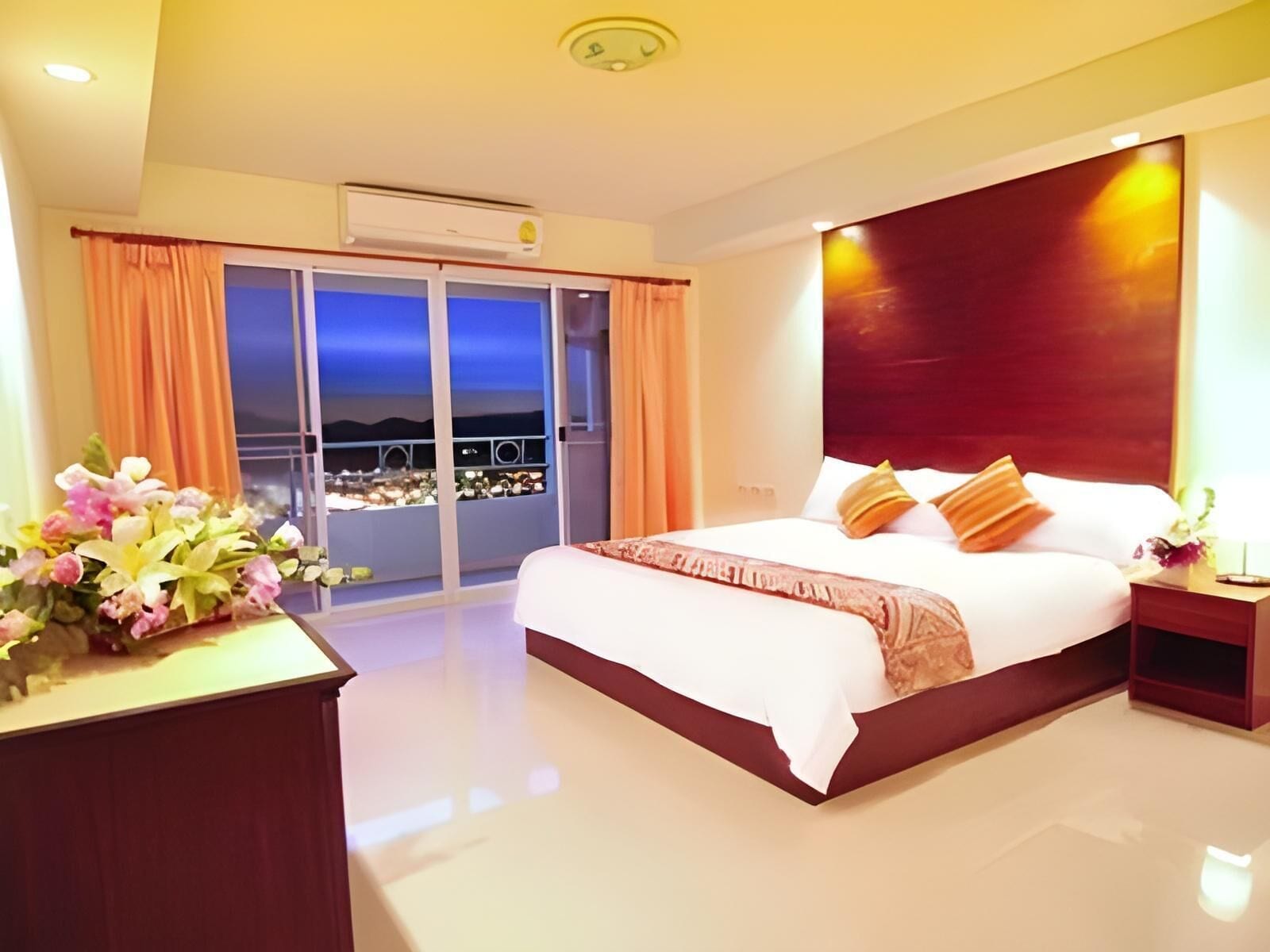A hotel room with a large bed, a nightstand, a desk with a lamp, and a view of the city at night through the balcony doors.
