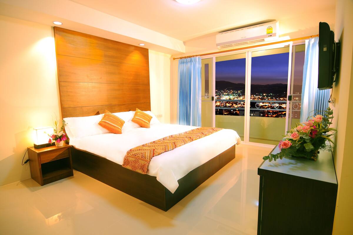 A hotel room with a large bed, a nightstand with a lamp, a TV mounted on the wall, and a view of a cityscape through the window.