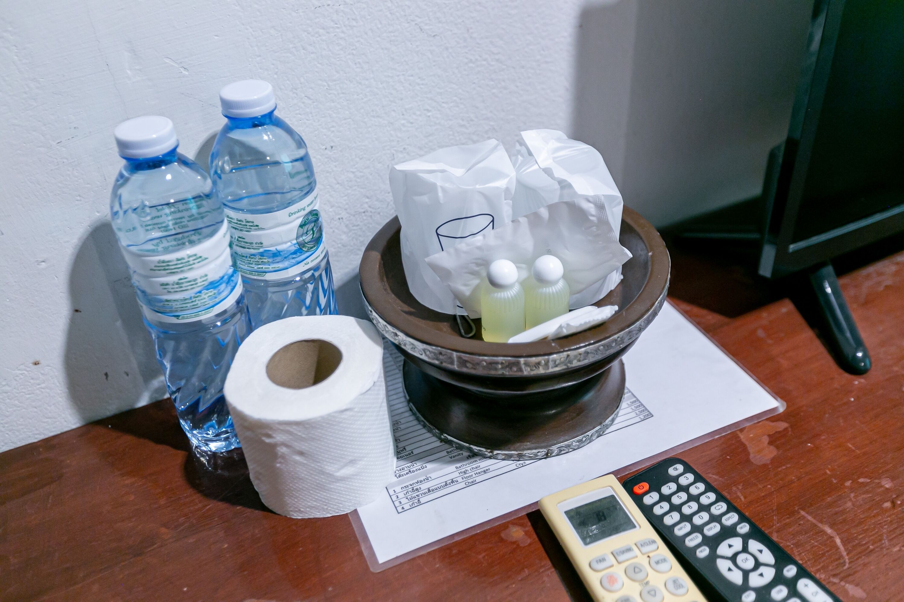 A table with bottled water, a roll of toilet paper, a remote control, and a bowl with tissues and a tissue box.