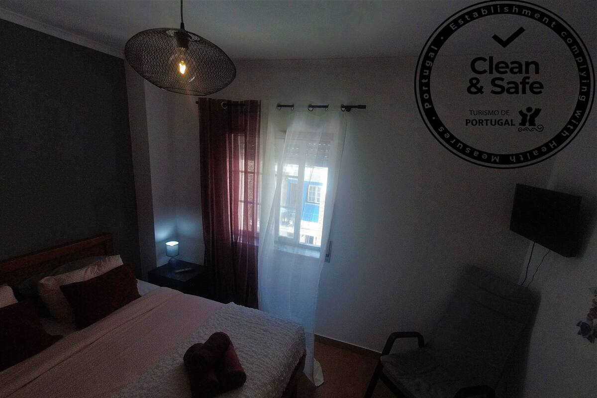 Habitación de hotel con cama, lámpara de noche, una silla, televisor montado en la pared y ventana con cortinas.