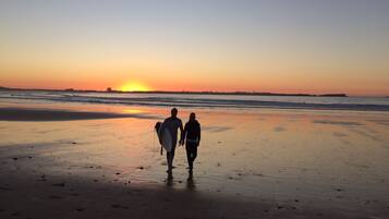 A beach at sunset with two people walking and carrying surfboards.