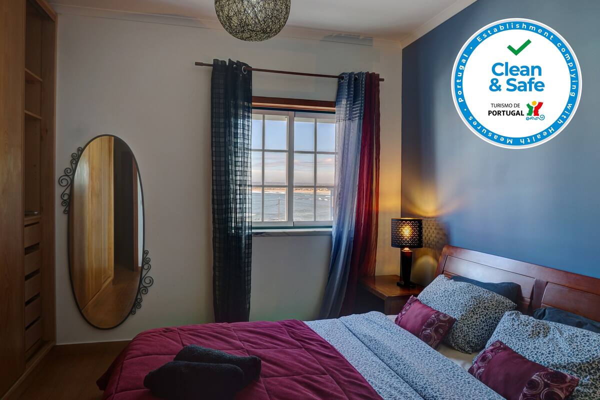 Un dormitorio con cama, mesita de noche, lámpara, ventana con cortinas, espejo y armario.