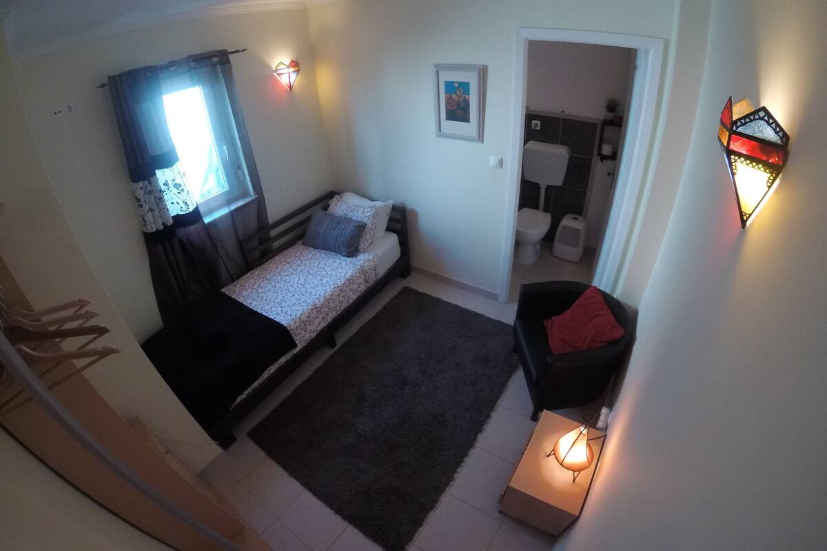 Un dormitorio pequeño y funcional con una cama individual, una mesita de noche, una lámpara, una ventana con cortinas y un baño visible a través de una puerta abierta.