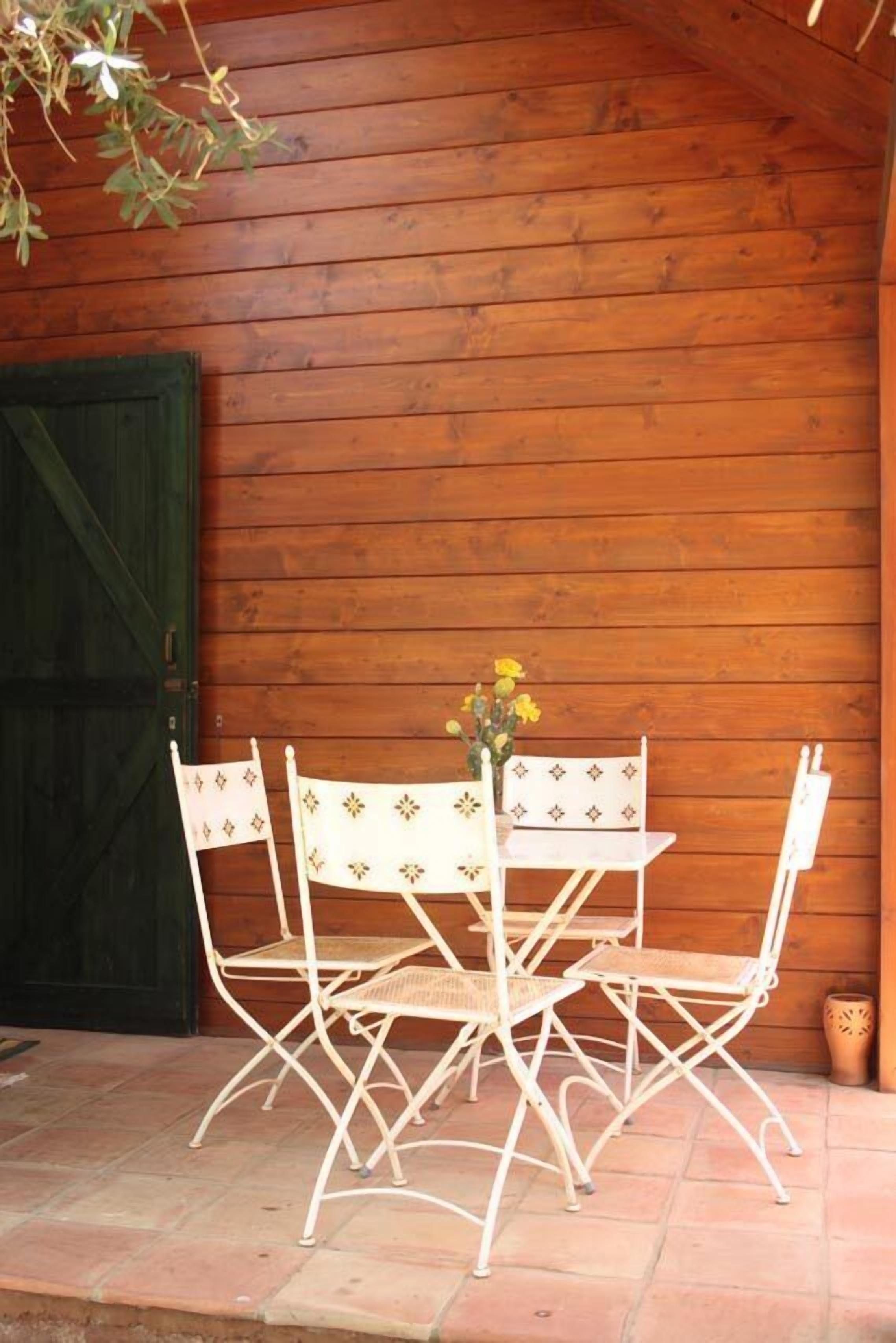 Un mur extérieur en bois, avec une porte verte et un espace patio comprenant une table et des chaises en métal blanc.