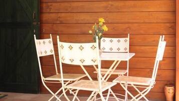 Un mur extérieur en bois, avec une porte verte et un espace patio comprenant une table et des chaises en métal blanc.