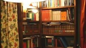Una habitación con una estantería llena de libros, un escritorio con papeles y una lámpara, y una ventana con cortinas.