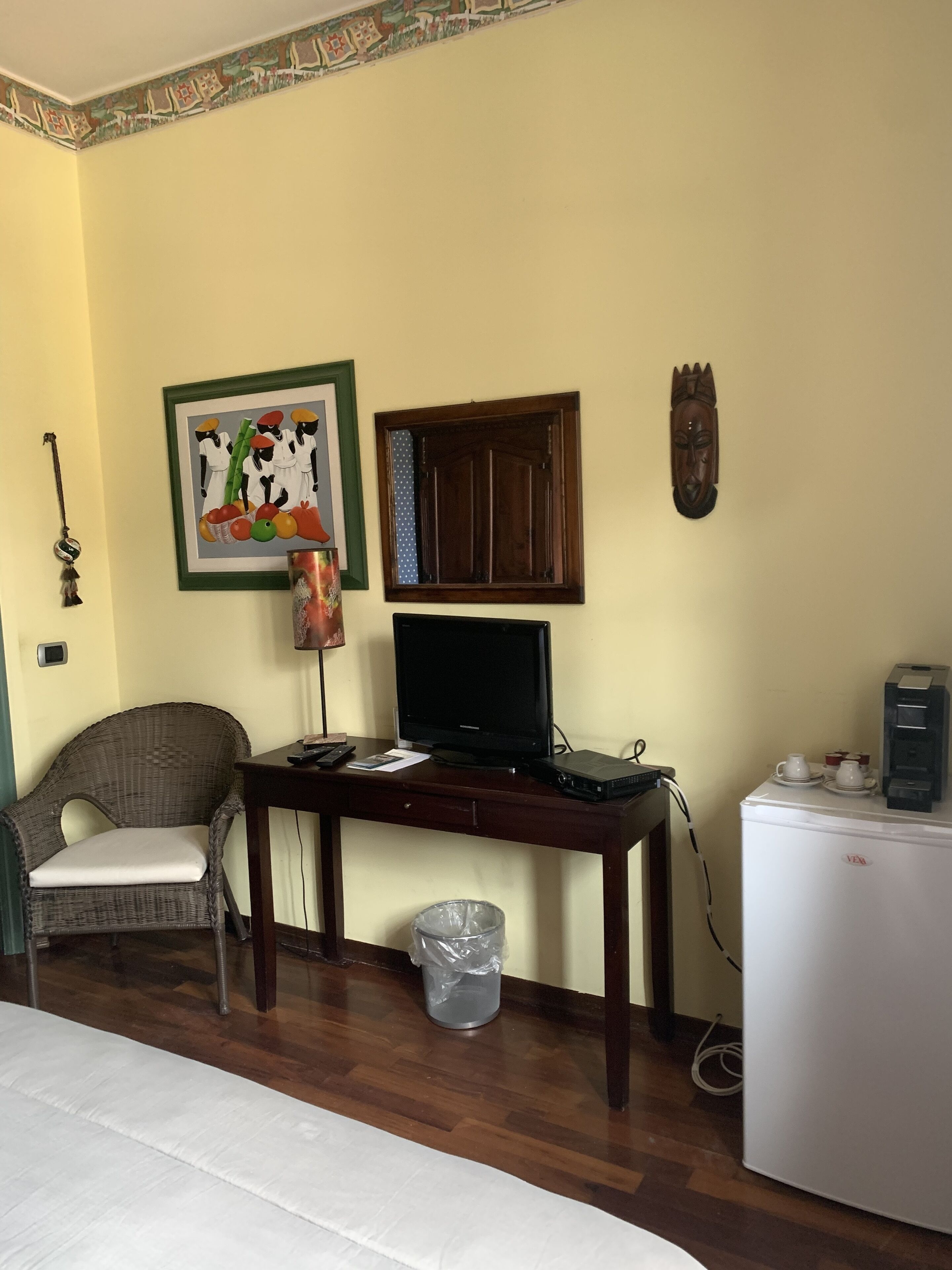 A room with a wooden desk, a television, a small refrigerator, a wicker chair, a trash bin, and wall decorations including a framed picture and a wooden mask.