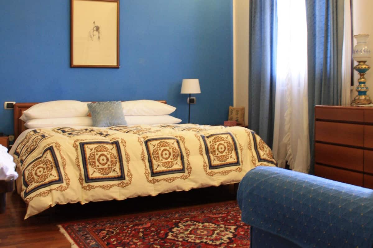 A bedroom with a bed, a blue wall, a wooden floor, a blue armchair, a dresser, and a lamp.
