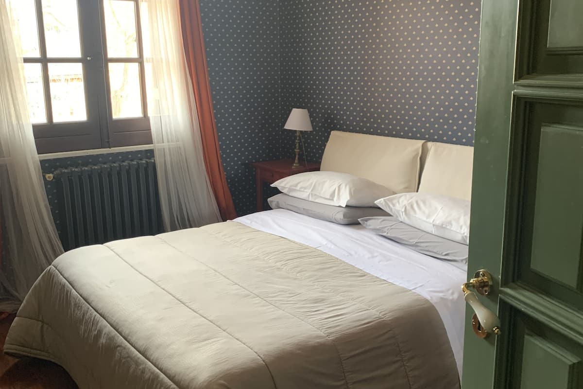 A neatly made bed with a beige comforter and pillows, a bedside table with a lamp, and a window with curtains.