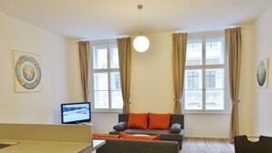 A modern living room with a grey sofa, orange cushions, a flat-screen TV, and large windows with curtains. - Luxury Oldtown Apartments (Vienna)