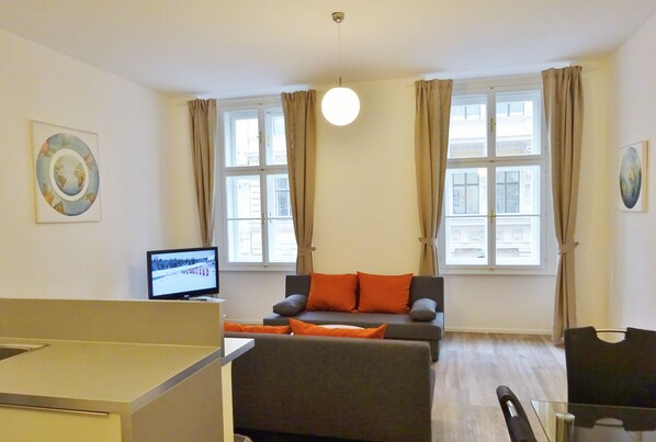A modern living room with a grey sofa, orange cushions, a flat-screen TV, and large windows with curtains.