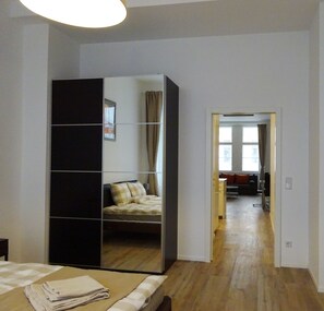 A bedroom with a large mirror wardrobe, a bed with checkered bedding, and a view into the living area through an open door.