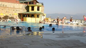 Zona de piscina con gente nadando y tomando el sol, una torre de salvavidas y vistas a montañas al fondo.