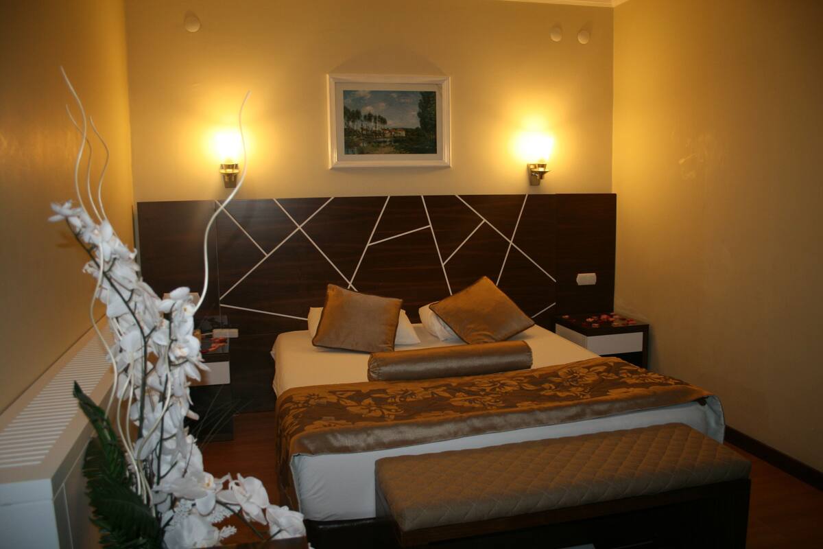 Una habitación de hotel con cama, mesitas de noche, un cuadro en la pared y una planta decorativa.