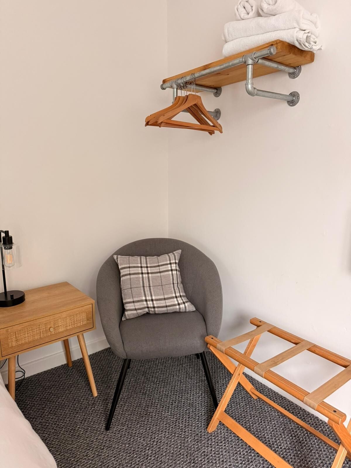 A room with a grey armchair, a small wooden side table with a lamp, a grey carpet, a wooden floor lamp, and a metal towel rack with hangers.