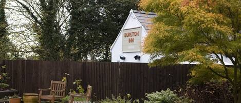 A white building with a sign reading "Burlton Inn" in a garden setting with a wooden fence, potted plants, and a bench.