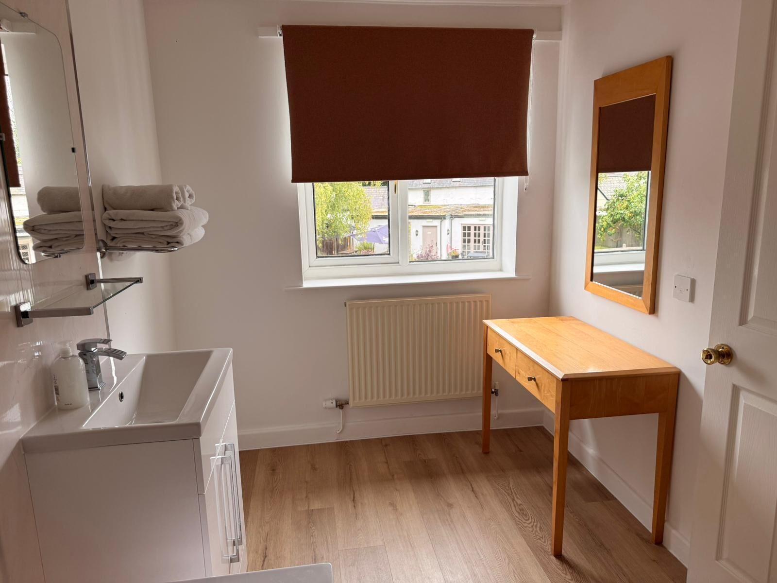 Een kleine, schone badkamer met een witte wastafel, spiegel en handdoekhouder. Er is een raam met een bruin zonnescherm, een houten bankje en een radiator.