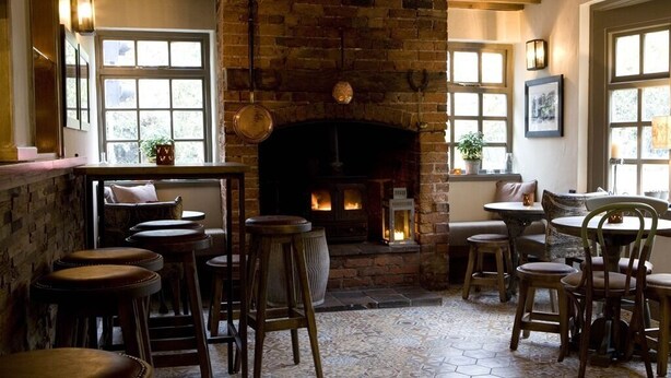 A cozy pub with a brick fireplace, wooden beams, hexagonal patterned floor tiles, and wooden bar stools.