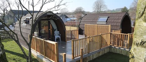 A small, enclosed wooden structure with a deck and a window, surrounded by a wooden fence and a grassy area.