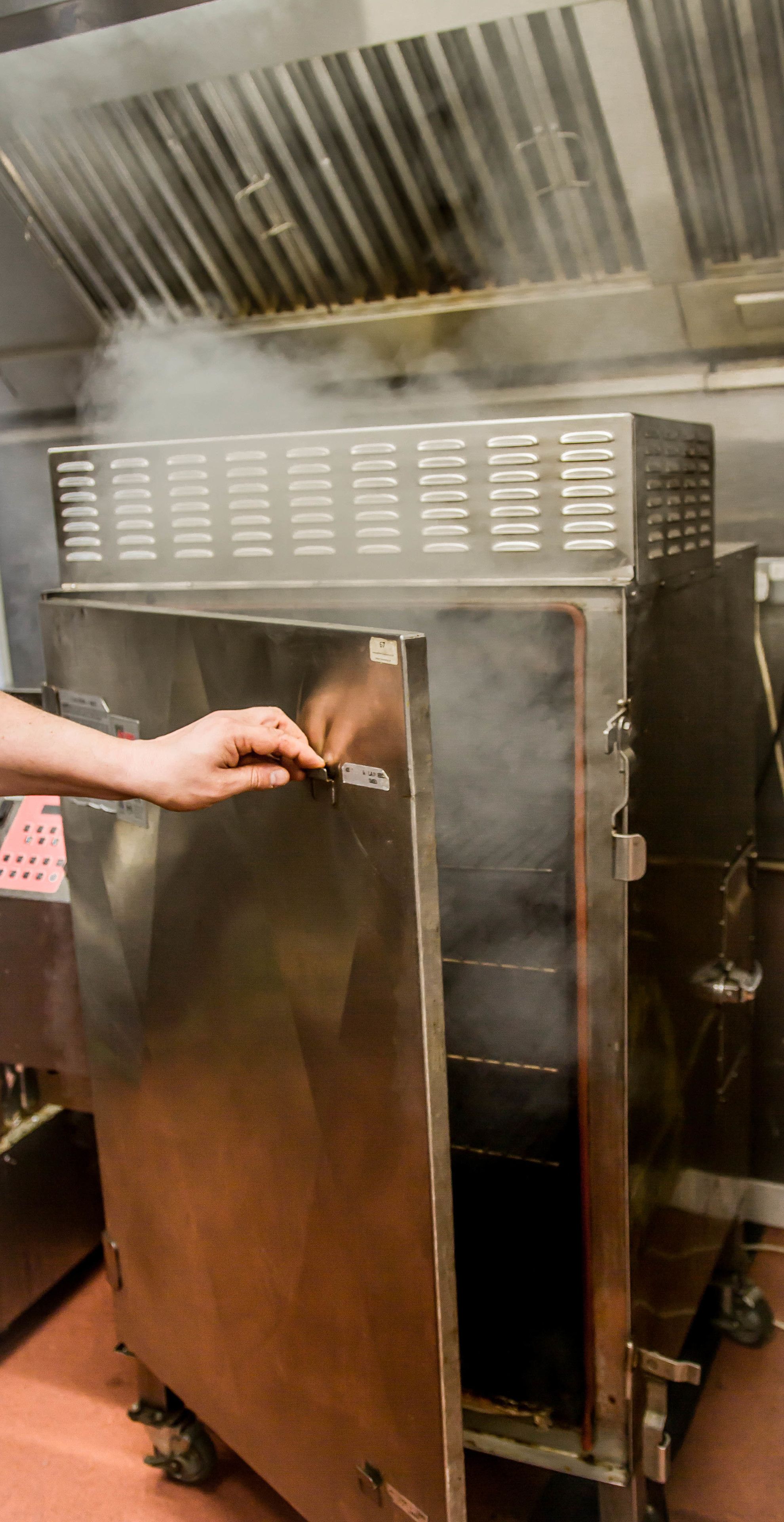 A person opening a large industrial oven door, with steam visible inside.