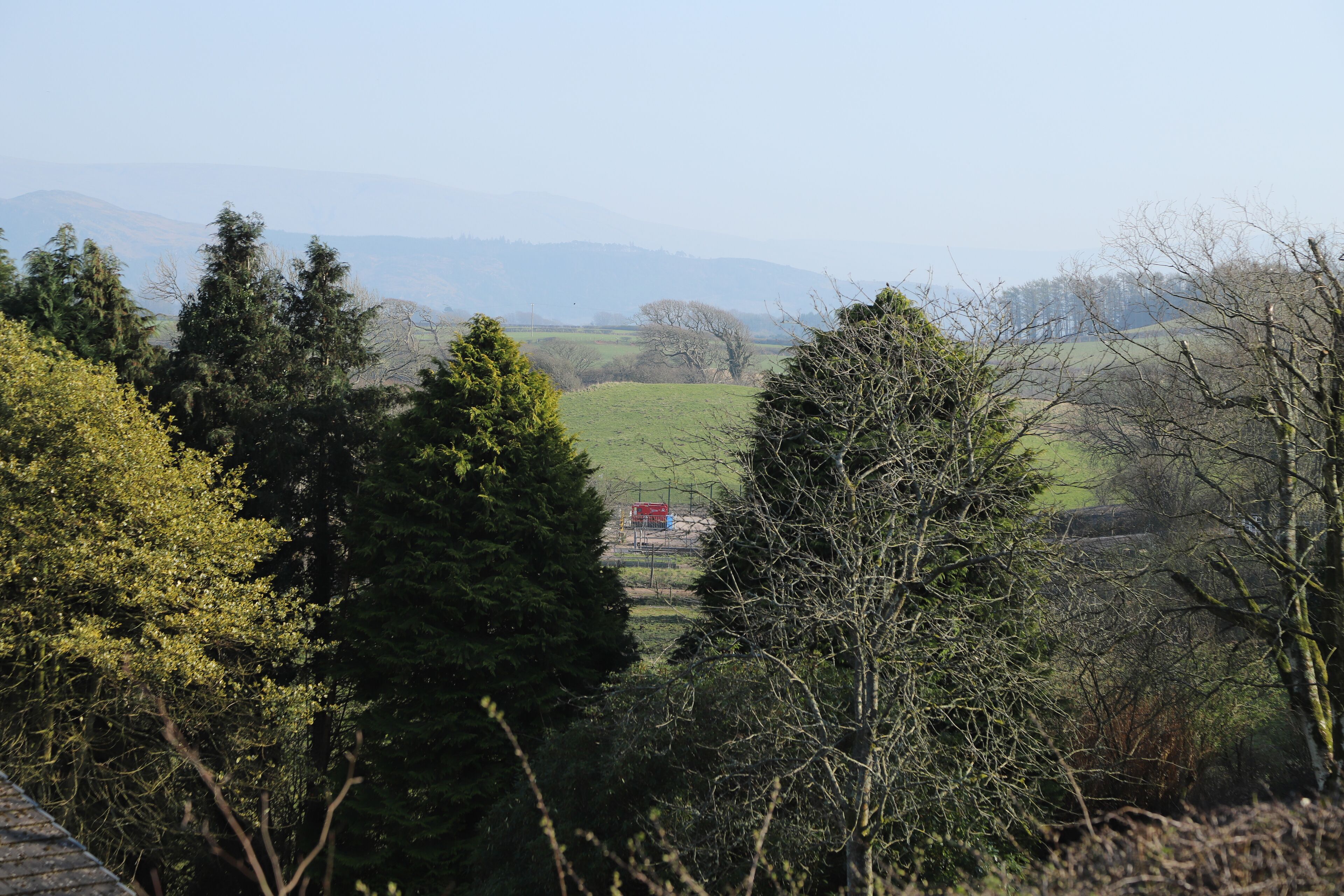 Un paysage rural avec une vue sur des montagnes lointaines, des arbres denses et un petit bâtiment rouge situé au milieu du paysage.