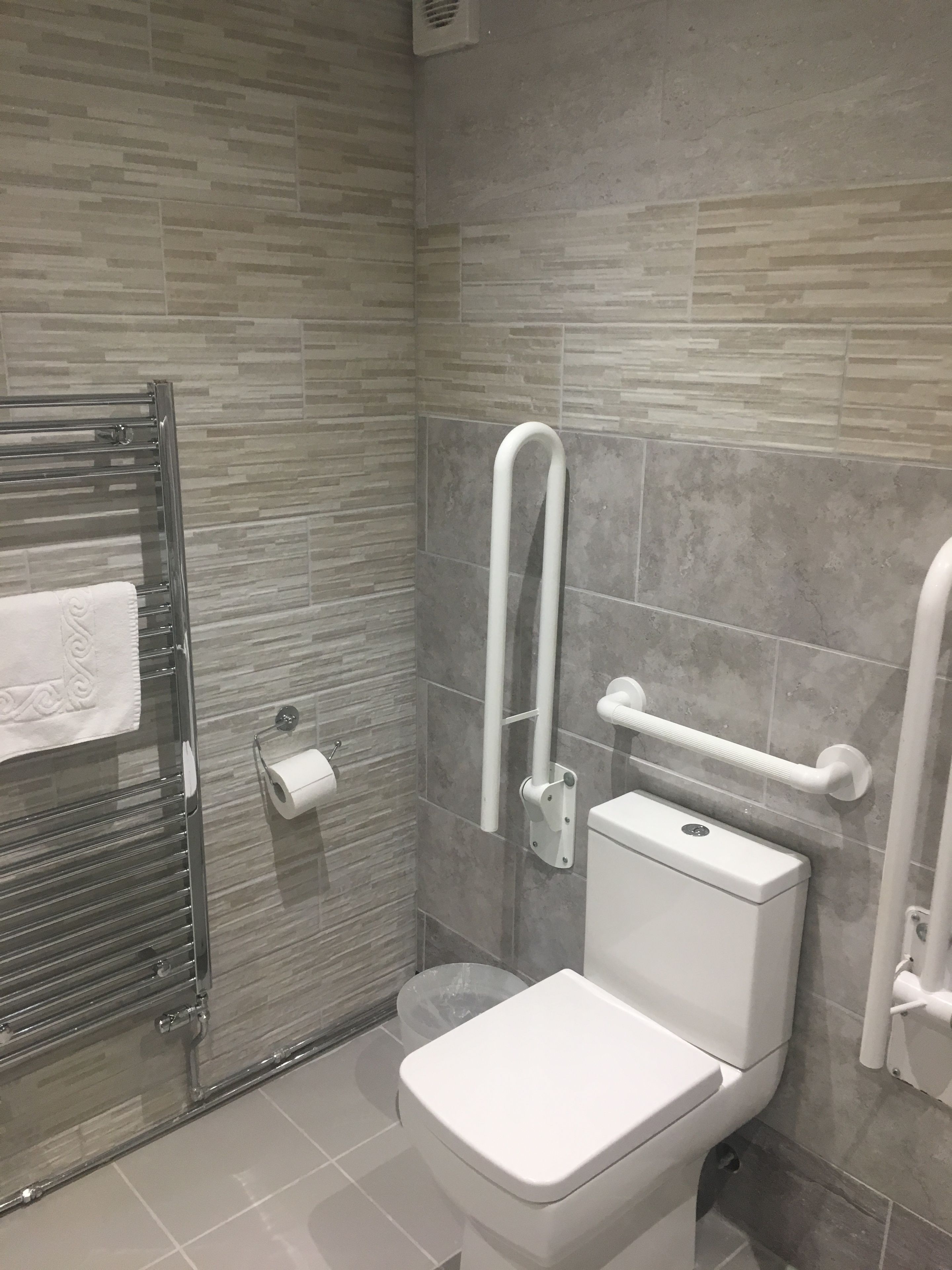 A modern bathroom with a toilet, grab bars, a towel rack, and a trash bin.